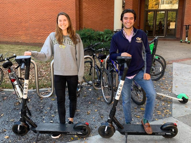 Two individuals standing with their scooters in front of the Instructional Center on Georgia Tech campus in front of a bike rack