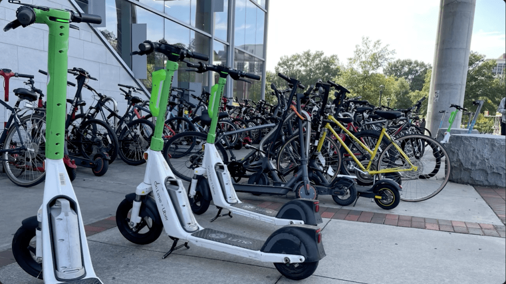 A plethora of scooters (both tap to pay Lime branded and personally owned) sitting at the North area micrmobility parking of the Clough Undergraduate Learning Commons. There are also many bikes locked in the background.