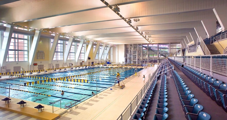 Interior view of the McAuley Aquatic Center inside the Georgia Tech Campus Recreation Center. The Olympic-sized swimming pool is divided into multiple lanes with blue and gold lane dividers. A few swimmers are in the water, and lifeguards stand nearby. Large windows line one side, flooding the space with daylight.