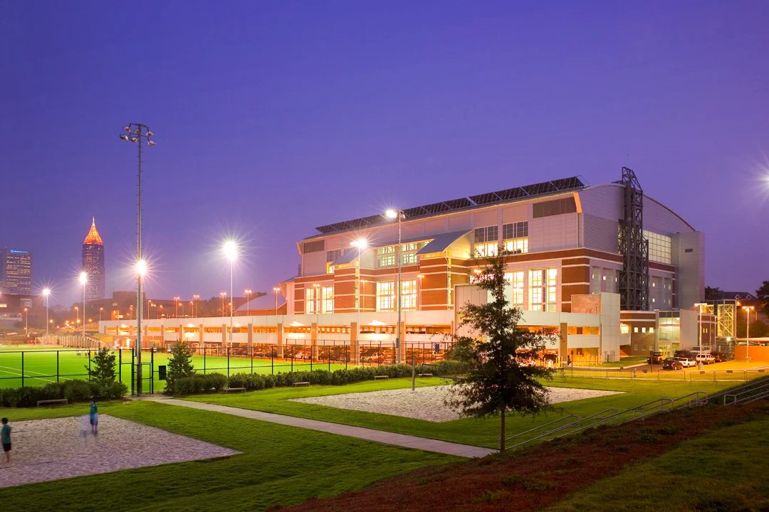 Exterior view of the Georgia Tech Campus Recreation Center at dusk, illuminated by tall floodlights that brighten the surrounding athletic fields. The building’s glass and brick façade glows warmly against the purple-blue evening sky. In the foreground, students are seen walking or playing near sand volleyball courts and grassy areas, with the Atlanta skyline faintly visible in the background.