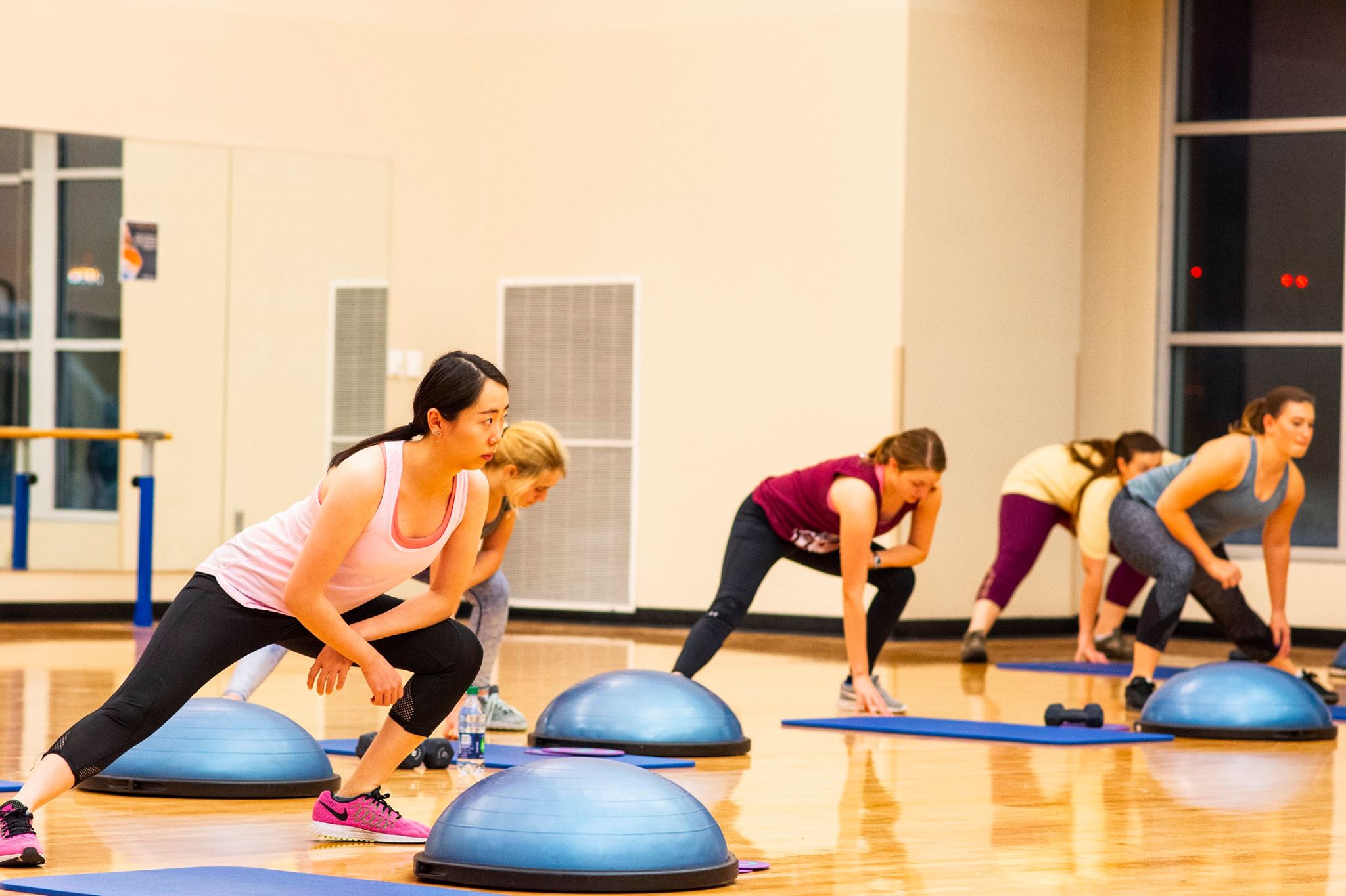 A group of students participating in a fitness class inside one of the CRC’s exercise studios. The participants, dressed in athletic clothing, are performing side lunges on blue mats with half-dome balance trainers in front of them. The polished wooden floor and mirrored wall reflect the bright lighting, while large windows line the back wall, giving the space an open and inviting look.