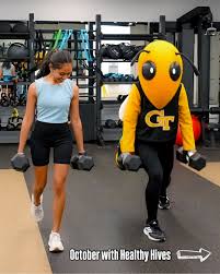 Inside the CRC’s strength training area, a Georgia Tech student in athletic gear performs dumbbell lunges beside the university’s mascot, Buzz, dressed in the iconic yellow jacket costume. The two are positioned on a clean gym floor surrounded by weight racks, mirrors, and exercise equipment, promoting fitness and school spirit.