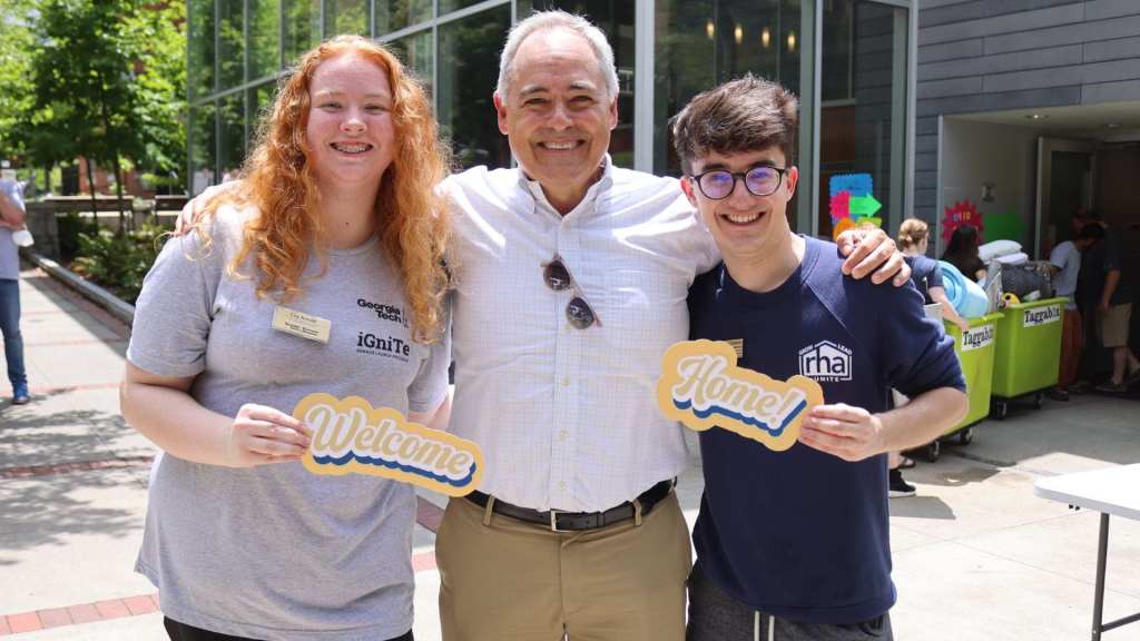President Cabrera stands outdoors with two smiling incoming freshmen in front of a campus building. The student on the left holds a sign that says "Welcome," and the student on the right holds a sign that says "Home!"