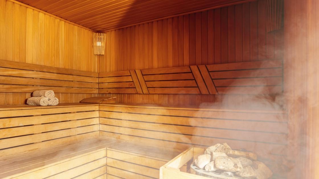 Interior view of a modern wooden sauna located inside the Georgia Tech CRC. The walls, ceiling, and benches are made of smooth, light-colored wood, with a small heater in the corner filled with hot stones producing visible steam. Rolled towels rest on the bench, and soft ambient light glows from a wall-mounted lamp, creating a calm, warm, and relaxing atmosphere.