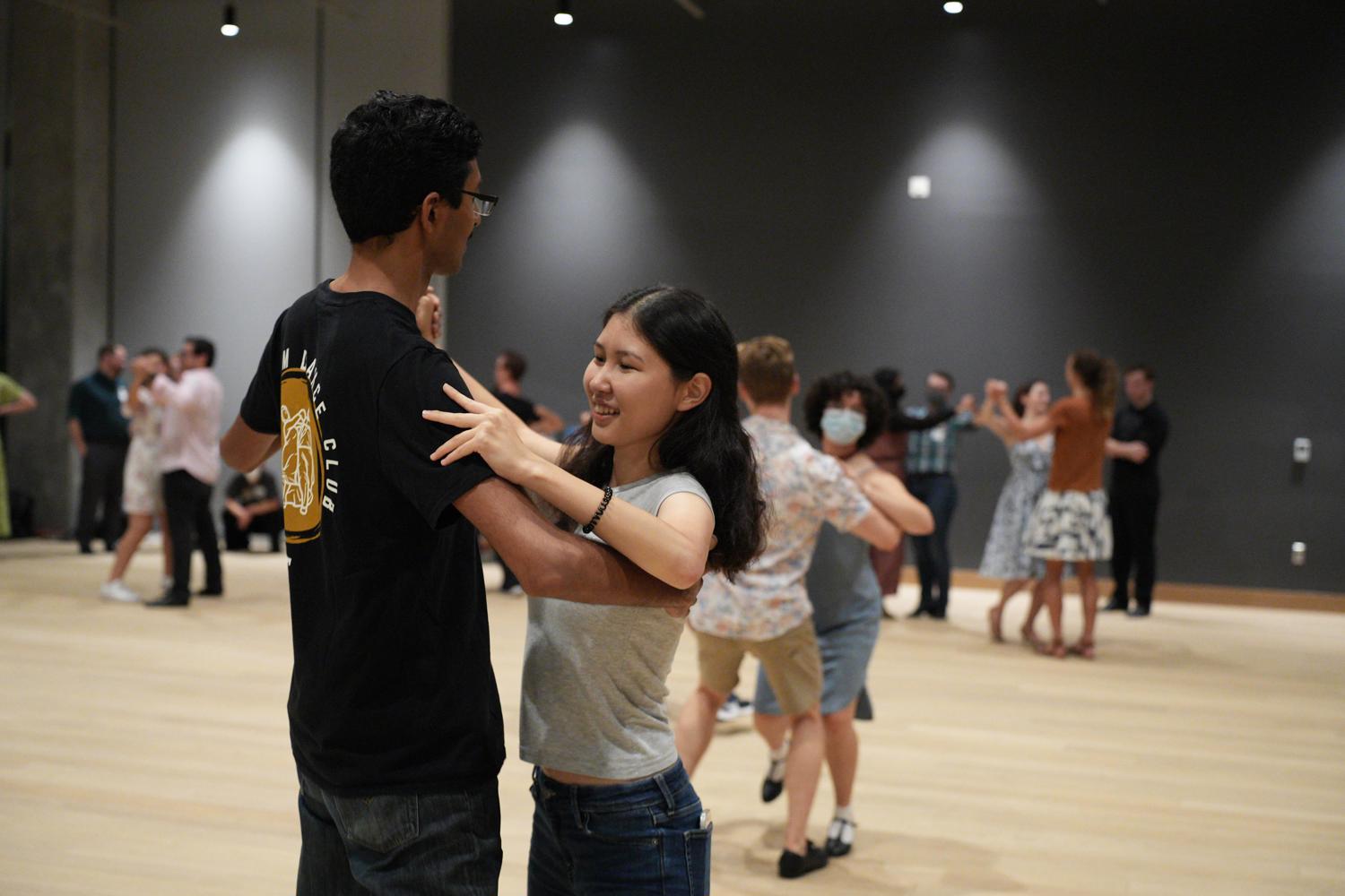 Students dancing with partners in a large indoor studio while other couples practice in the background.
