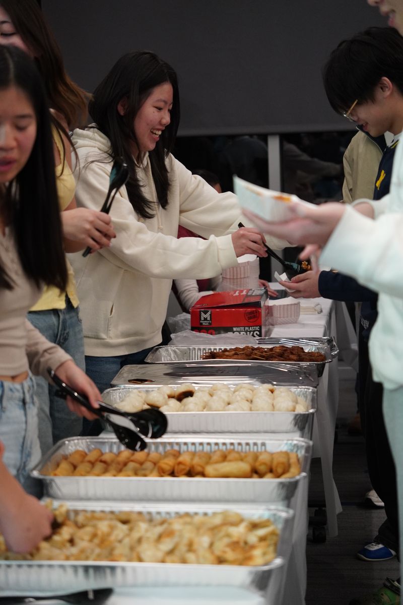 Students serve themselves food from a buffet table lined with trays of dishes at an indoor gathering.