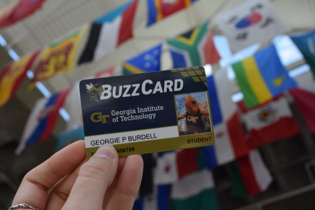 Hand holding a Georgia Tech BuzzCard student ID in the foreground, with colorful international flags blurred in the background.