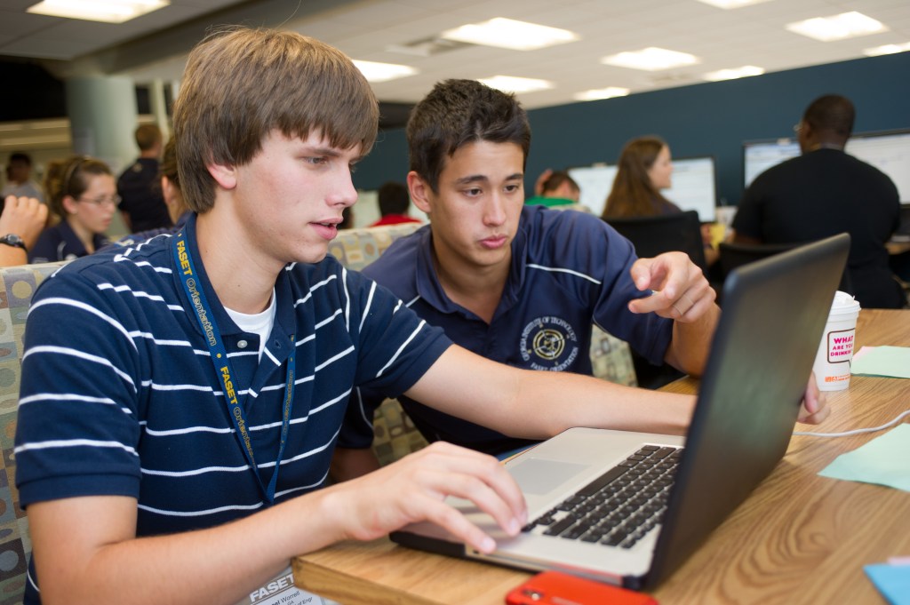 A student sitting at a table with a laptop while a teaching assistant points at the screen and helps explain course material during office hours. Other students are visible working in the background.