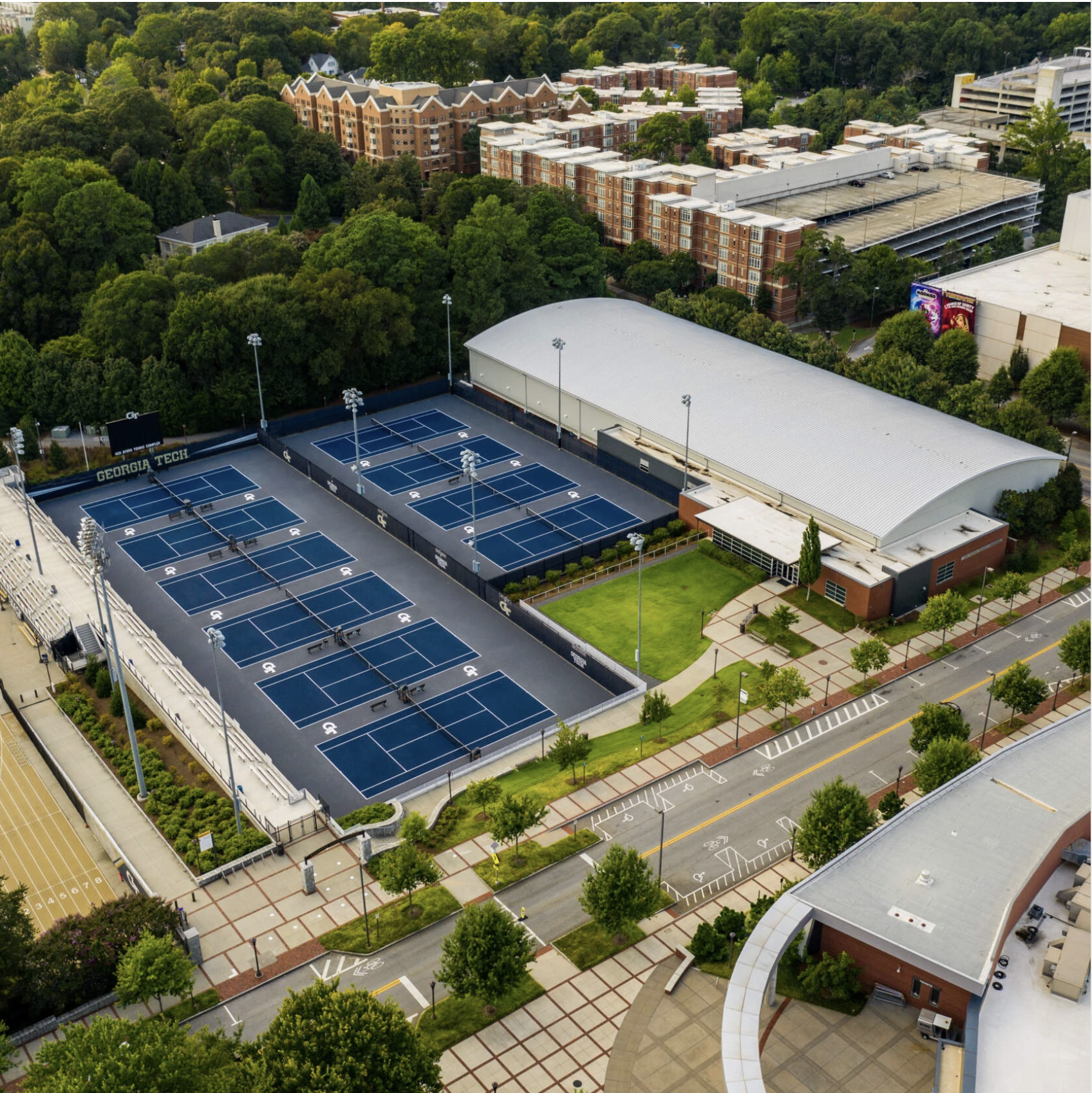Aerial view of Georgia Tech’s tennis complex with multiple blue outdoor courts, adjacent indoor facility, surrounding trees, and nearby campus buildings.
