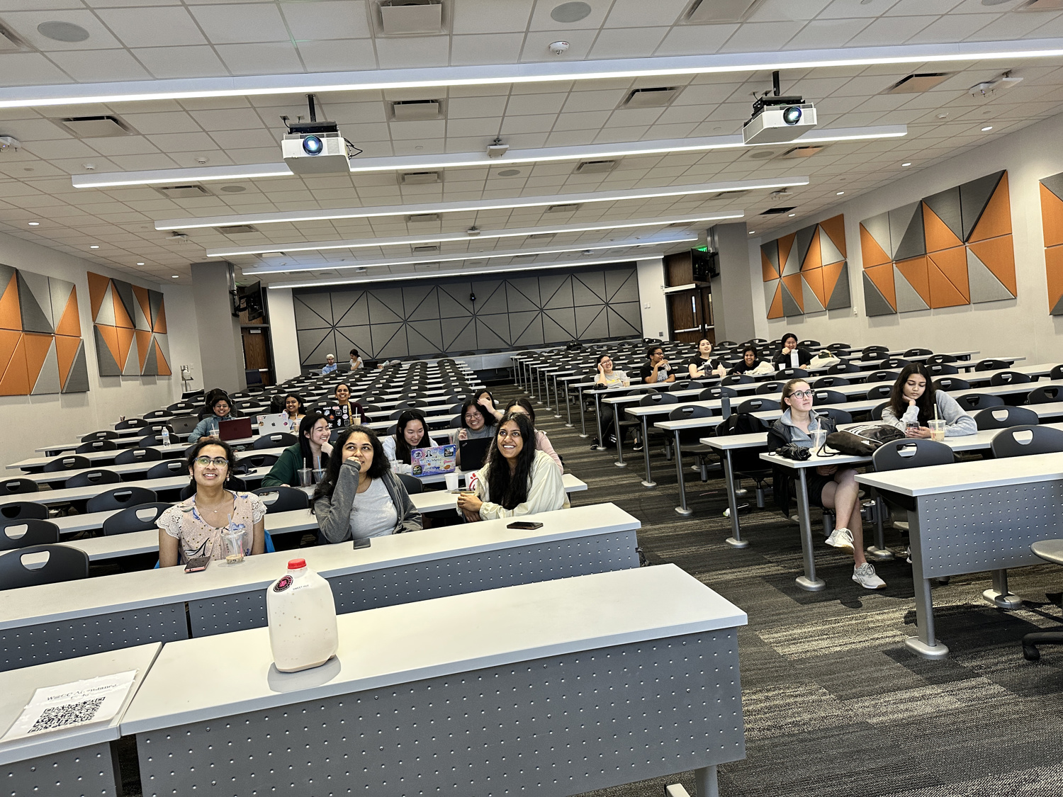 Attendees seated throughout a large lecture hall during an intro meeting for Women @ College of Computing, with several students smiling toward the camera and others using laptops or talking at their tables.

