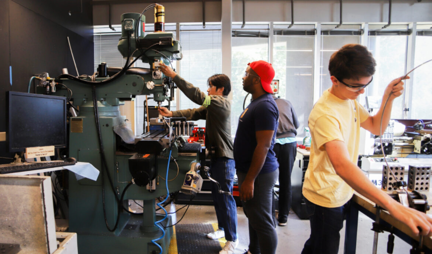 One student operating a vertical milling machine inside the Flowers Invention Studio, with one student in safety glasses works at a nearby workbench in the foreground.