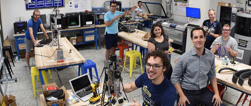 Smiling students in the Yang Aero Maker Space, surrounded by workbenches, 3D printers, laser cutters, and various hands-on engineering projects.