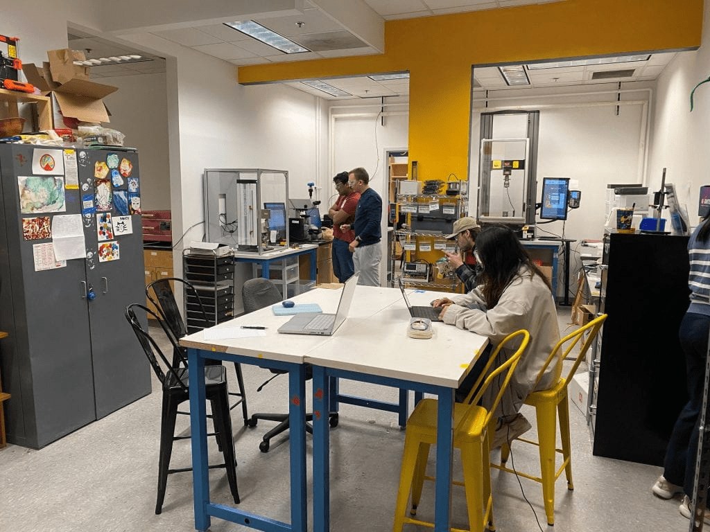 A student working at a table in the MILL with two other students in the background operating a testing machine.