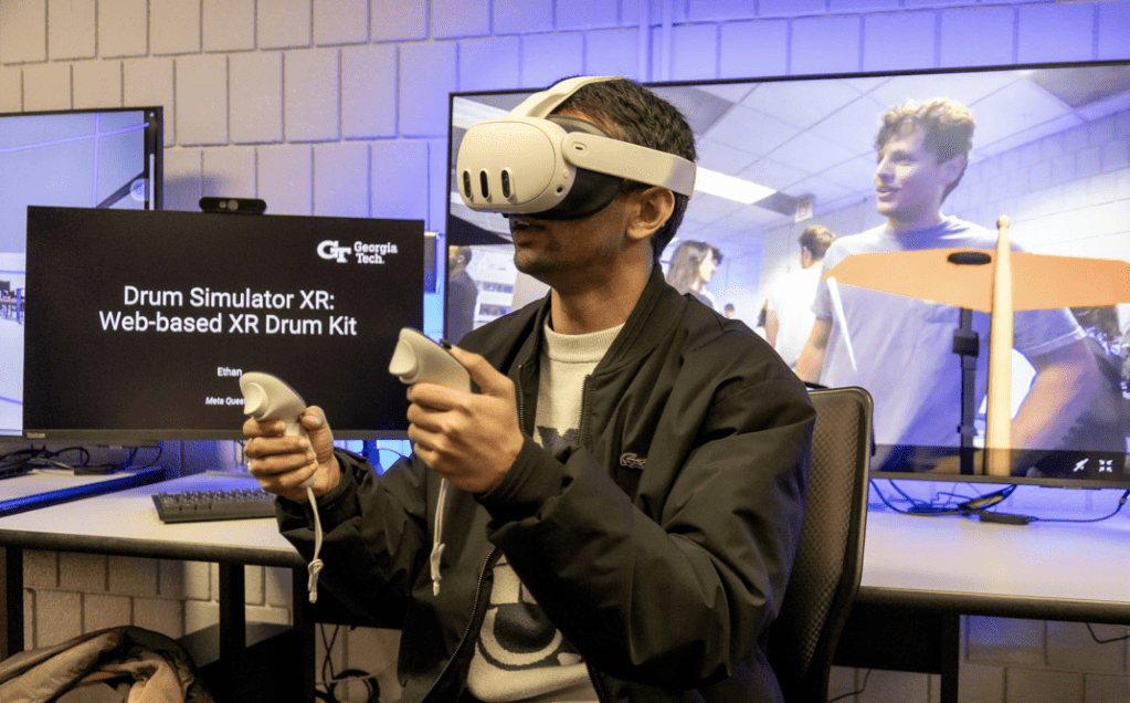 A student is seated in the XR makerspace utilizing the Meta Quest 3 to test a XR drum kit. Computer monitors behind him display information on what he is testing and the makerspace.
