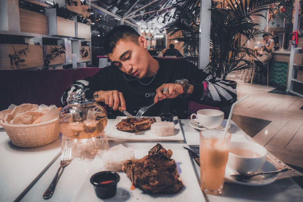 A young man in a black sweatshirt cuts into a grilled meat dish served with rice at a restaurant table. A glass teapot, a basket of crackers, a drink, and a second plate sit in the foreground.