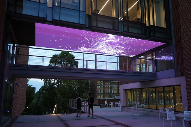 Two students walking through the breezeway between Georgia Tech's Price Gilbert Library and Crosland Tower beneath an illuminated ceiling.