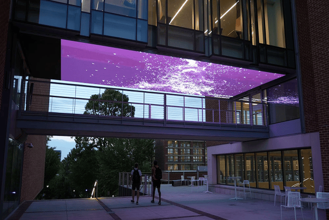 Exterior view of the breezeway between Price Gilbert and Crosland Tower at the Georgia Tech Library, showing two students walking under the pedestrian bridge and purple LED ceiling display.
