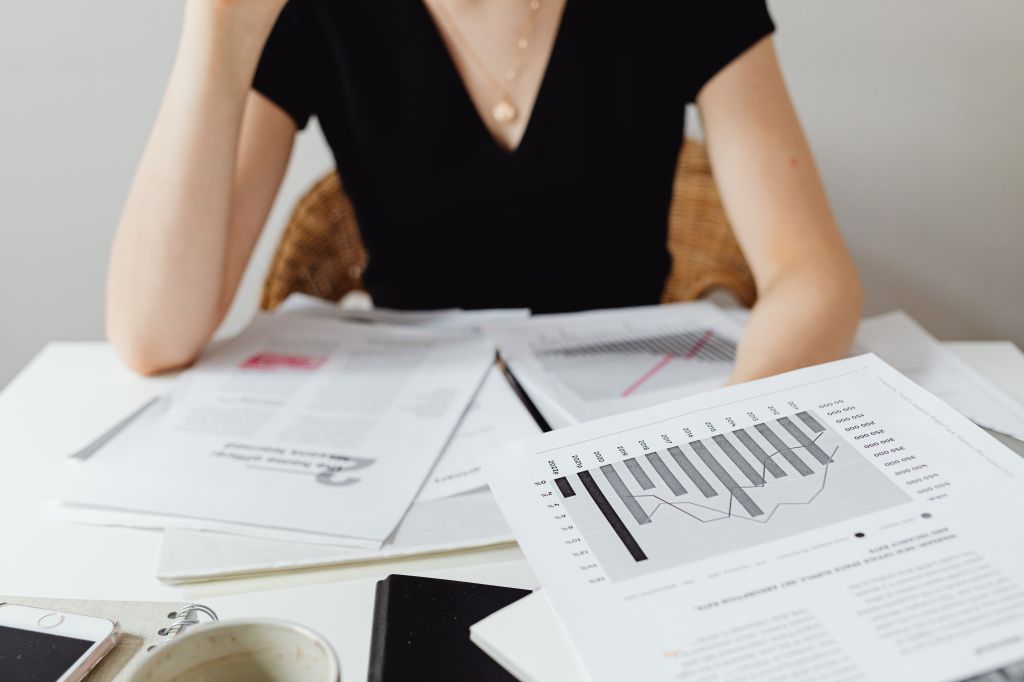 A woman sits at a desk reviewing financial documents, including printed reports, bar graphs, and line charts showing financial data and trends.