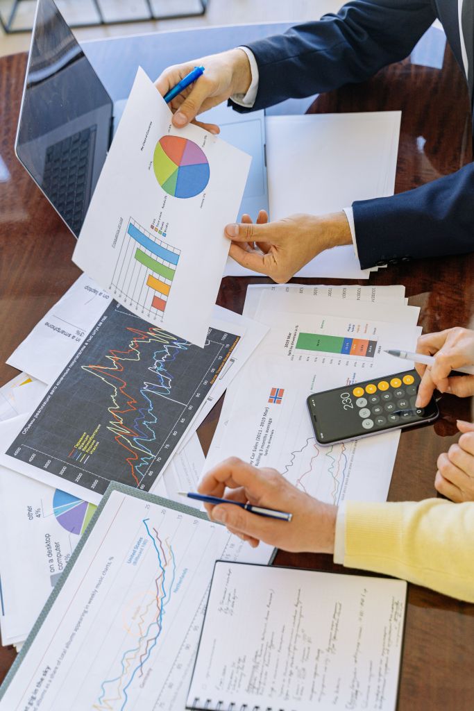 People reviewing financial reports around a table, including stock market charts, pie charts, and bar graphs, while using a calculator and laptop to analyze financial data.