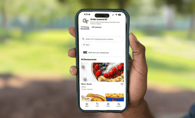 Phone screen held in one hand showing the Grubhub app’s “Order around GT” page. The GT Dining tab is selected, with a search bar, a campus card balance, and an All Restaurants list that begins with a Blenz Bowls listing and a photo of a berry bowl.