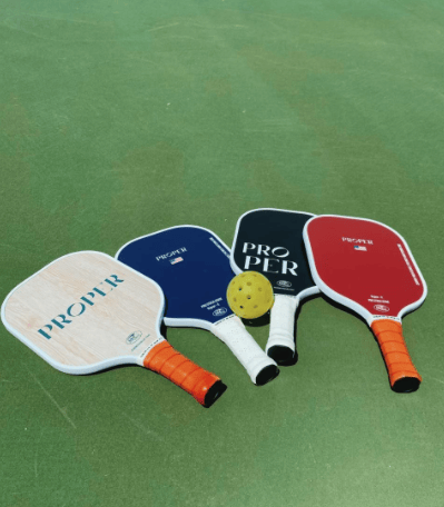 Four pickleball paddles in white, blue, black, and red with a yellow pickleball resting on top, on the floor of a green court