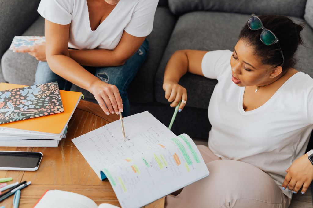 Two women seated beside a wooden coffee table collaborate on handwritten study notes. One woman points to an open notebook with a pencil while the other looks closely at the pages, holding a green pencil. Colorful highlighted notes, notebooks, folders, and writing supplies are spread across the table.