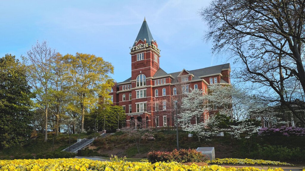 Georgia Tech’s Tech Tower, a red brick building with a tall pointed roof, surrounded by green trees, blooming white flowers, and landscaped gardens on a sunny day.
